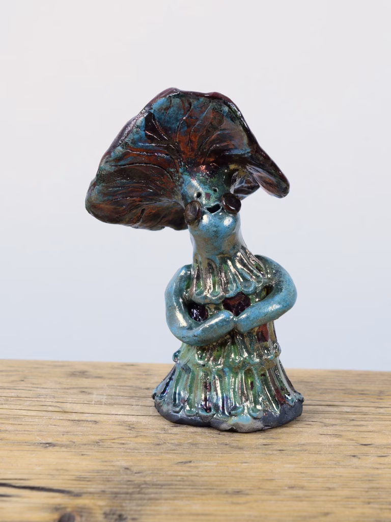 Prudence, a raku-fired ceramic mushroom lady with folded arms and a tilted cap in teal, copper and burgundy